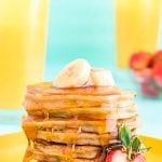 Close up photo of a stack of banana pancakes on a yellow plate topped with sliced banana and a strawberry on the side.