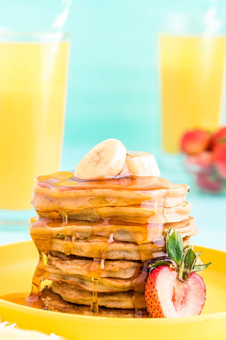 Close up photo of a stack of banana pancakes on a yellow plate topped with sliced banana and a strawberry on the side.