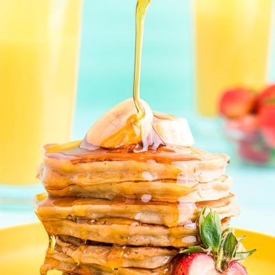 Close up photo of a stack of banana pancakes on a yellow plate with maple syrup being poured over the top.
