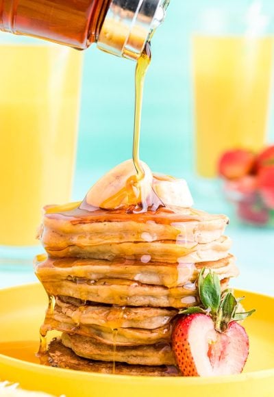 Close up photo of a stack of banana pancakes on a yellow plate with maple syrup being poured over the top.