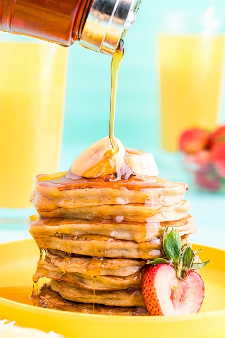 Close up photo of a stack of banana pancakes on a yellow plate with maple syrup being poured over the top.