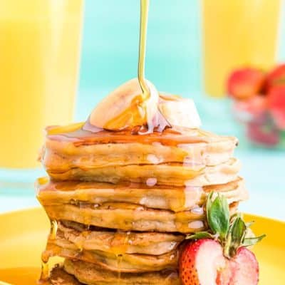Close up photo of a stack of banana pancakes on a yellow plate with maple syrup being poured over the top.