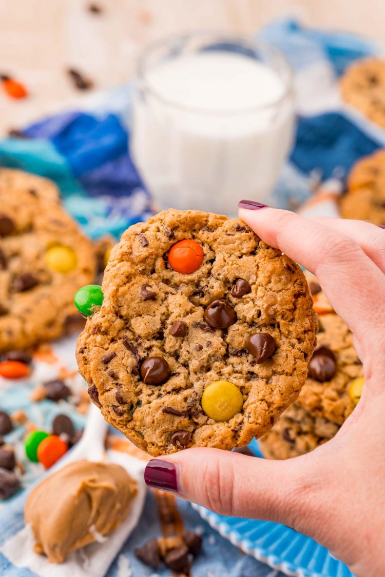 A woman's hand holding a monster cookie with a blue napkin and glass of milk in the background.