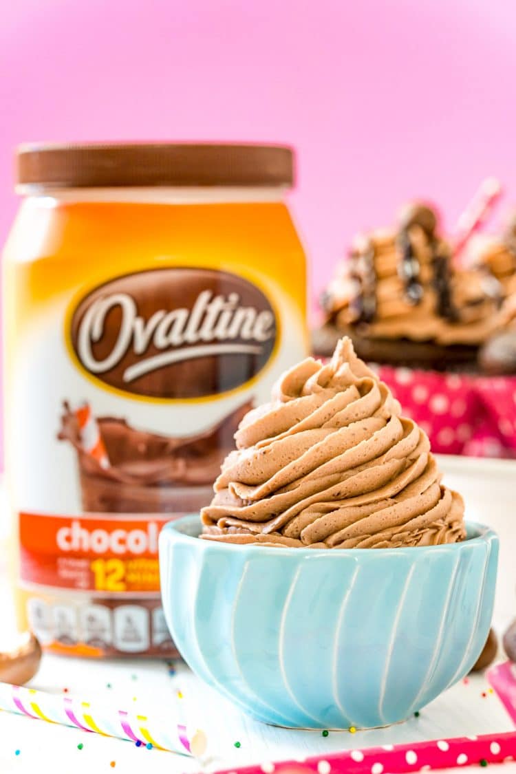 Small blue bowl filled with chocolate malt frosting with a container of Ovaltine and cupcakes in the background.