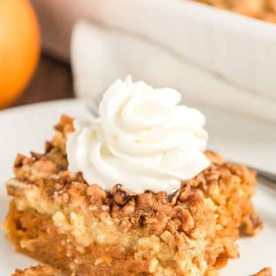 Close up photo of a slice of pumpkin dump cake on a white plate with pumpkin and more cake in background.