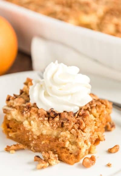 Close up photo of a slice of pumpkin dump cake on a white plate with pumpkin and more cake in background.