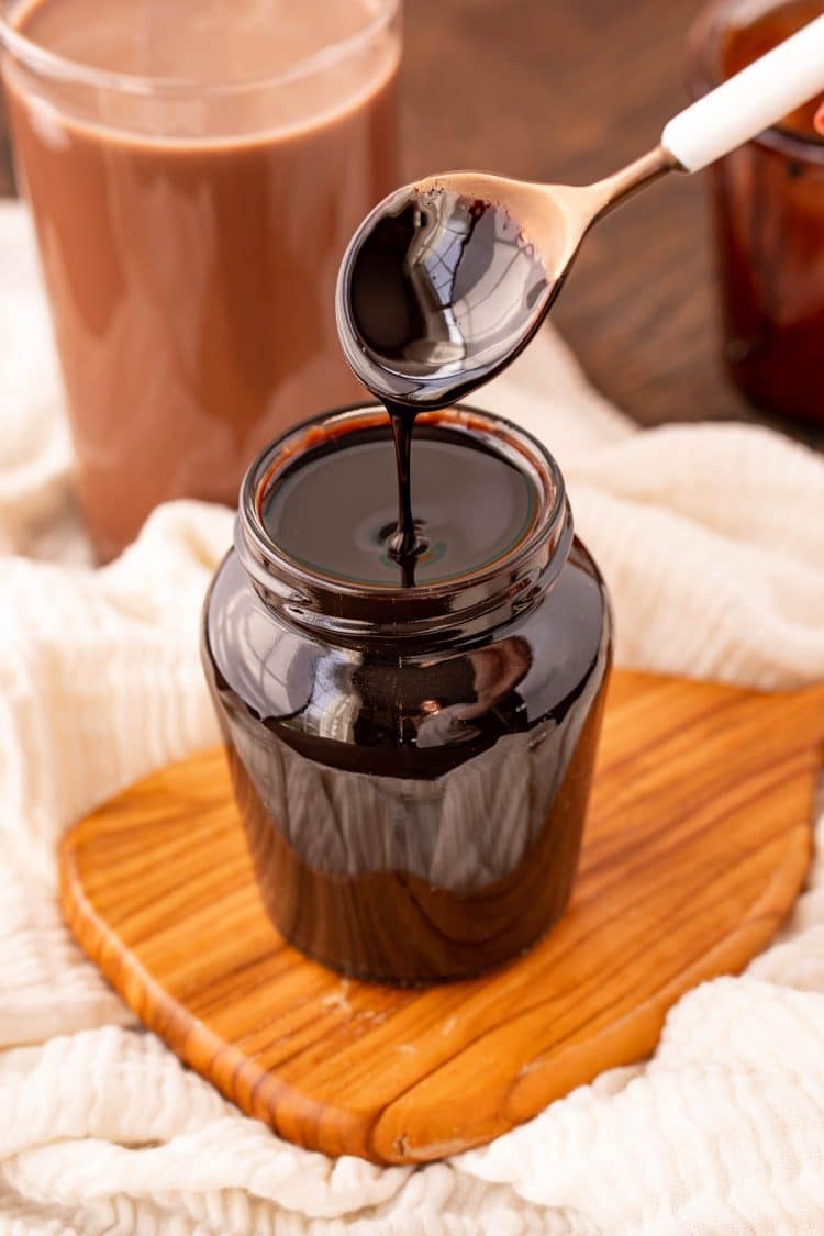 A spoon drizzling chocolate syrup into a jar full of it.