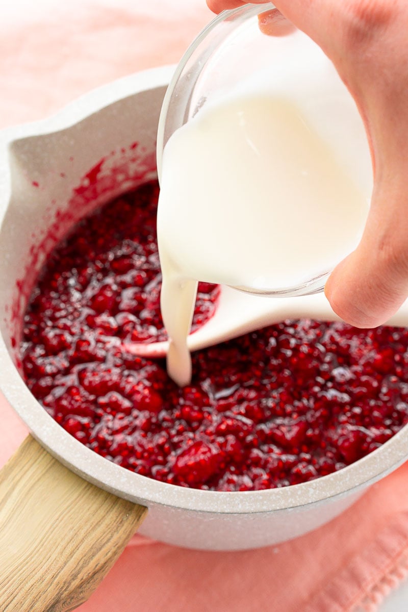 Cornstarch and water mixture being added to a pot of cooking raspberries.