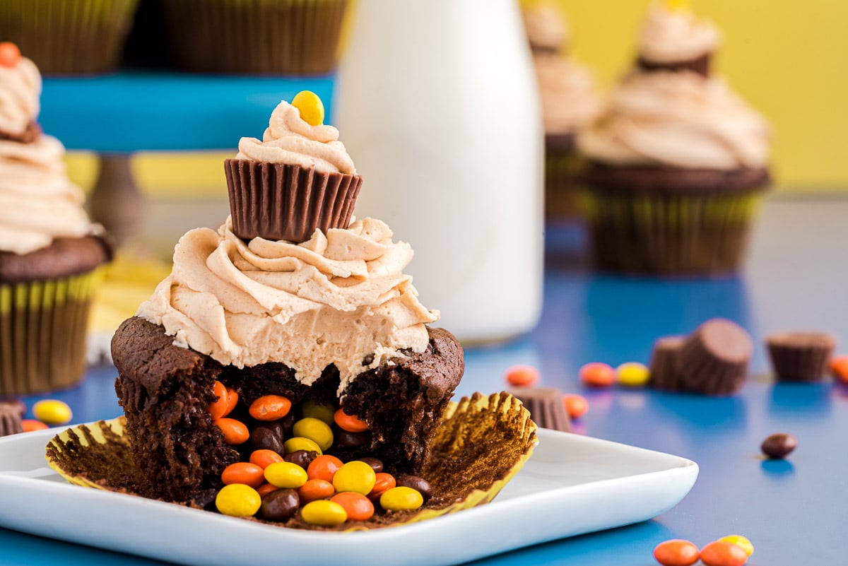 A chocolate cupcake with peanut butter frosting on a white plate with reese's pieces spilling out of the center where a bite has been taken.