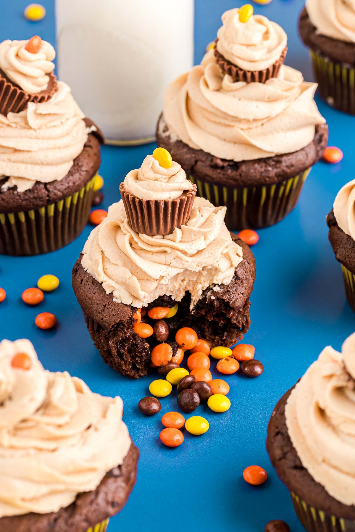 Close up photo of peanut butter chocolate cupcakes on a blue surface with a glass of milk and Reese's pieces scattered around.