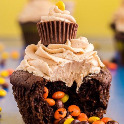 Close up photo of a peanut butter chocolate cupcake with mini Reese's pieces in the center falling out onto a blue surface.