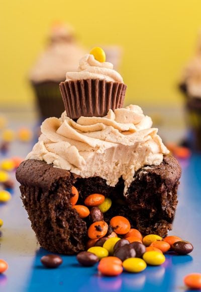 Close up photo of a peanut butter chocolate cupcake with mini Reese's pieces in the center falling out onto a blue surface.