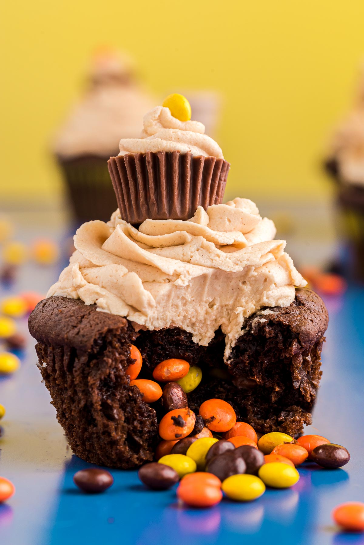 Close up photo of a peanut butter chocolate cupcake with mini Reese's pieces in the center falling out onto a blue surface.