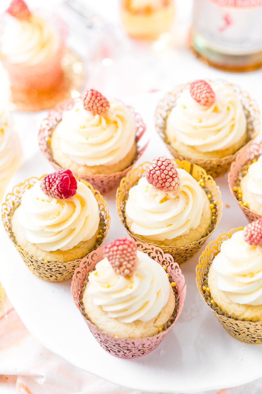 Champagne Cupcakes on a cake stand with a bottle of champagne and more cupcakes in the background.