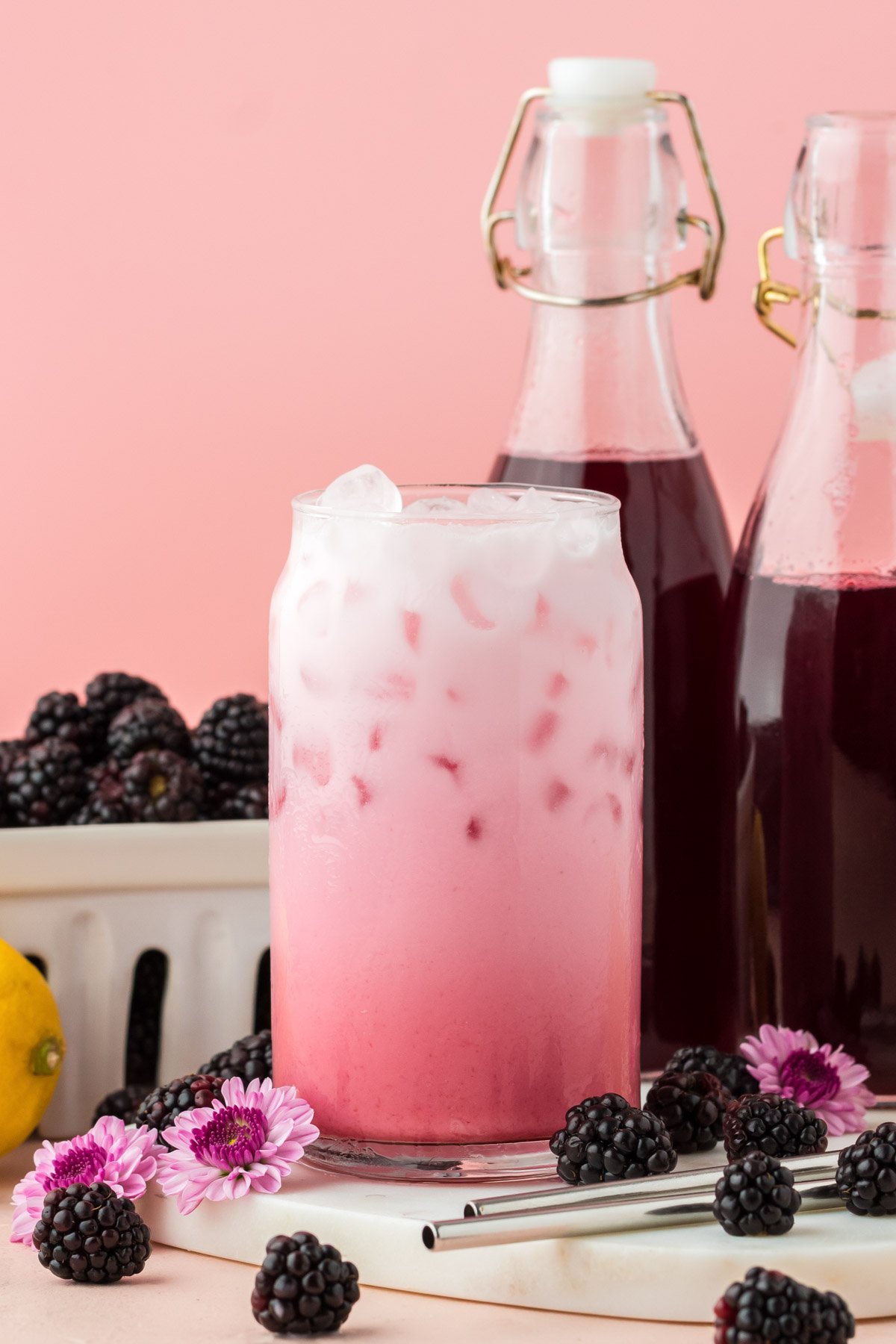 A glass of blackberry Italian cream soda on a white serving board with bottle of syrup and blackberries in the background.