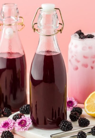 Close up photo of two glass bottles of blackberry simple syrup and a glass with a blackberry cream soda.