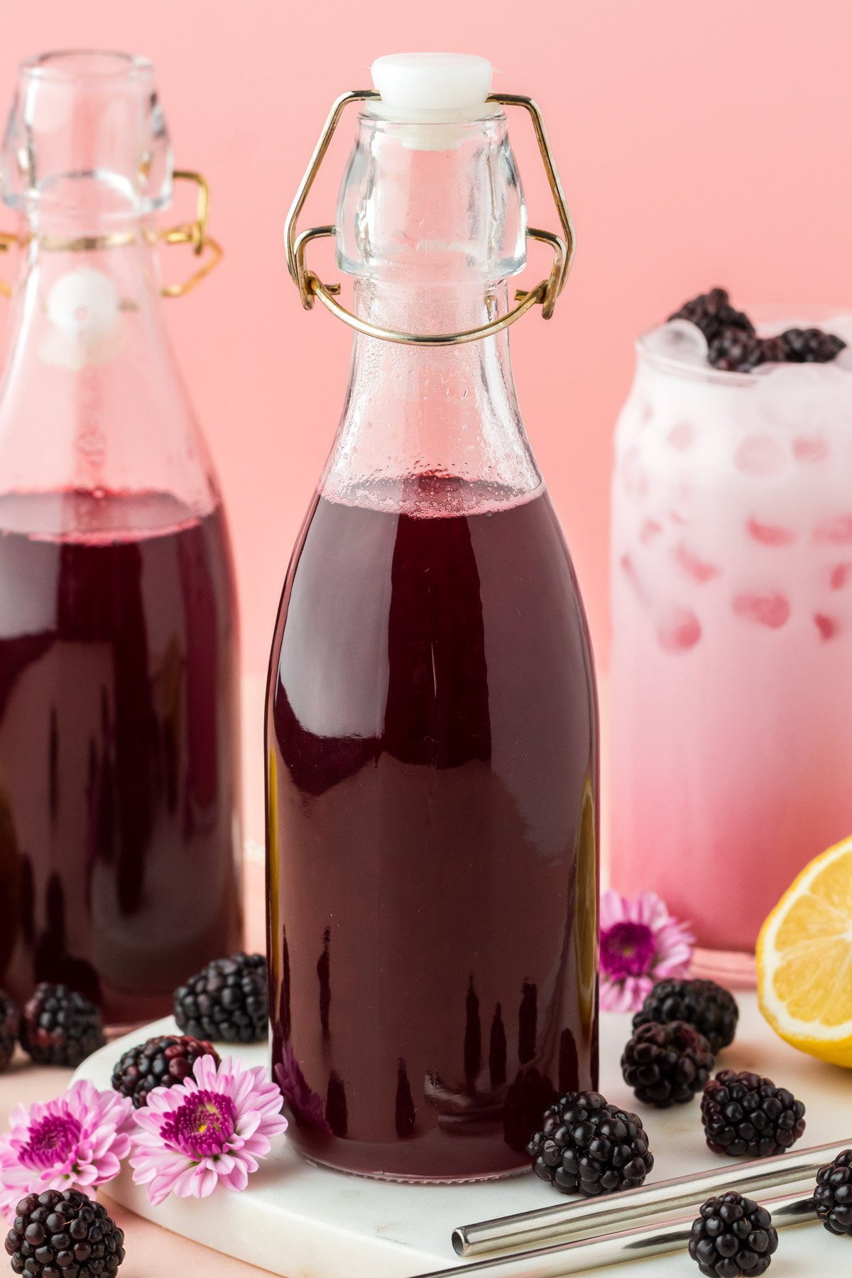 Close up photo of two glass bottles of blackberry simple syrup and a glass with a blackberry cream soda.