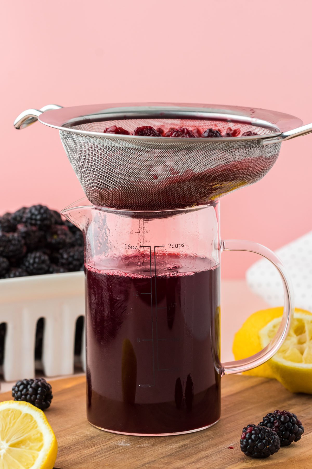 A fine mesh sieve straining blackberry simple syrup into a measuring cup.