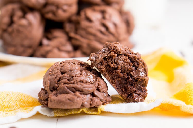 Two chocolate cookies on a yellow napkin in front of a plate of more cookies.