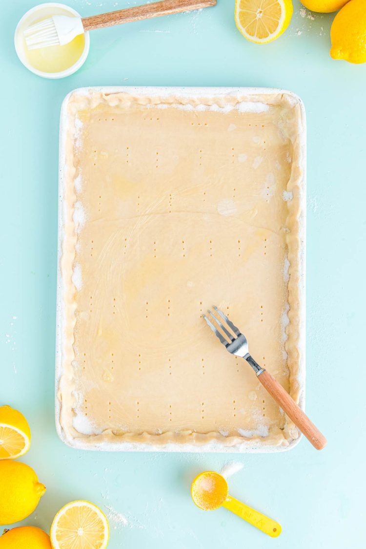 Pie crust in a jelly roll pan that has been pricked with a fork. lemons and a bowl of butter surround it.
