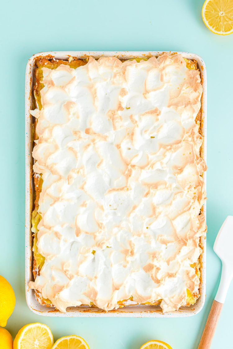 Overhead photo of a lemon meringue slab pie on a blue surface with lemons around it.