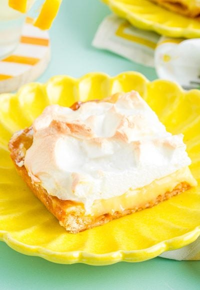 Close up photo of a slice of lemon meringue slab pie on a yellow plate on a blue surface.