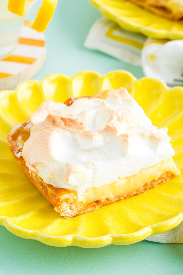 Close up photo of a slice of lemon meringue slab pie on a yellow plate on a blue surface.