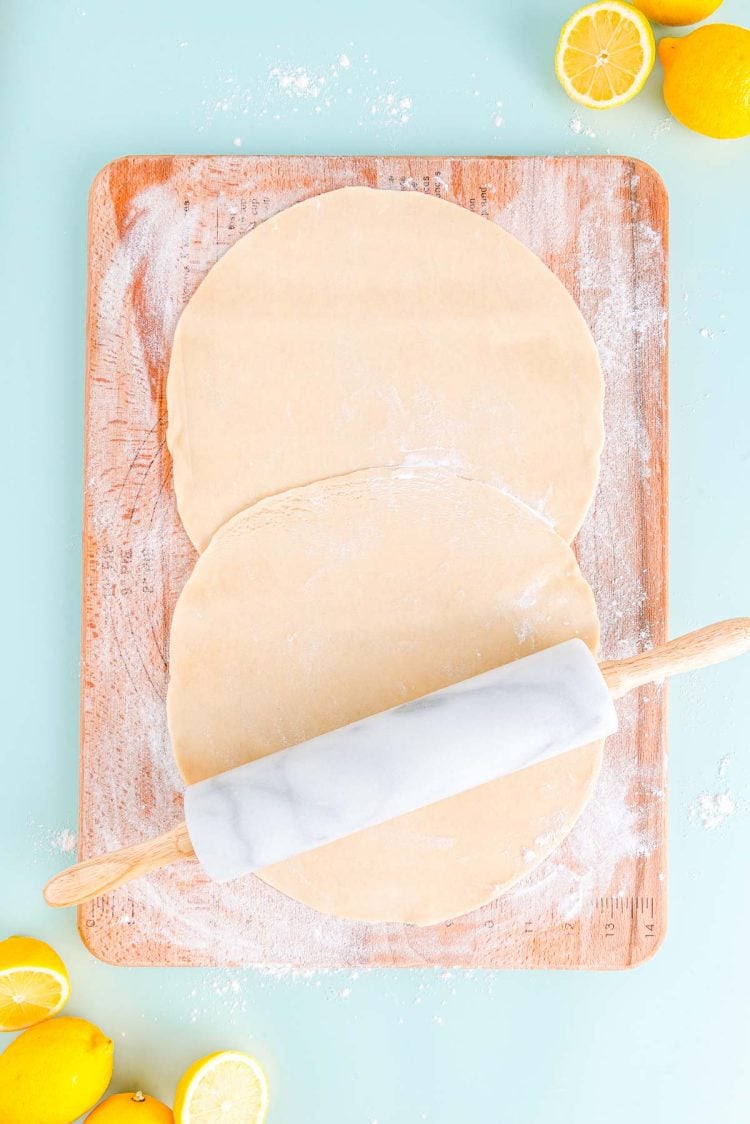 Pie crusts being rolled out on a cutting board on a blue surface.