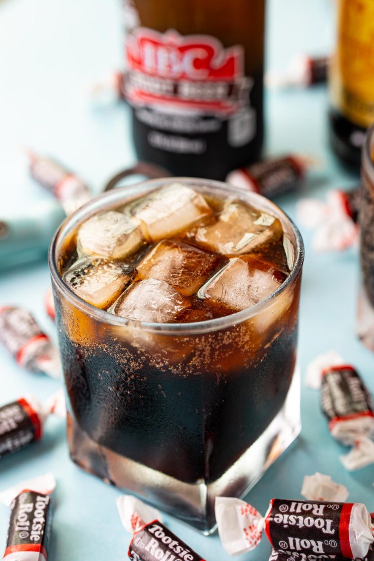 A glass filled with a tootsie roll cocktail on a blue surface surrounded by tootsie rolls.