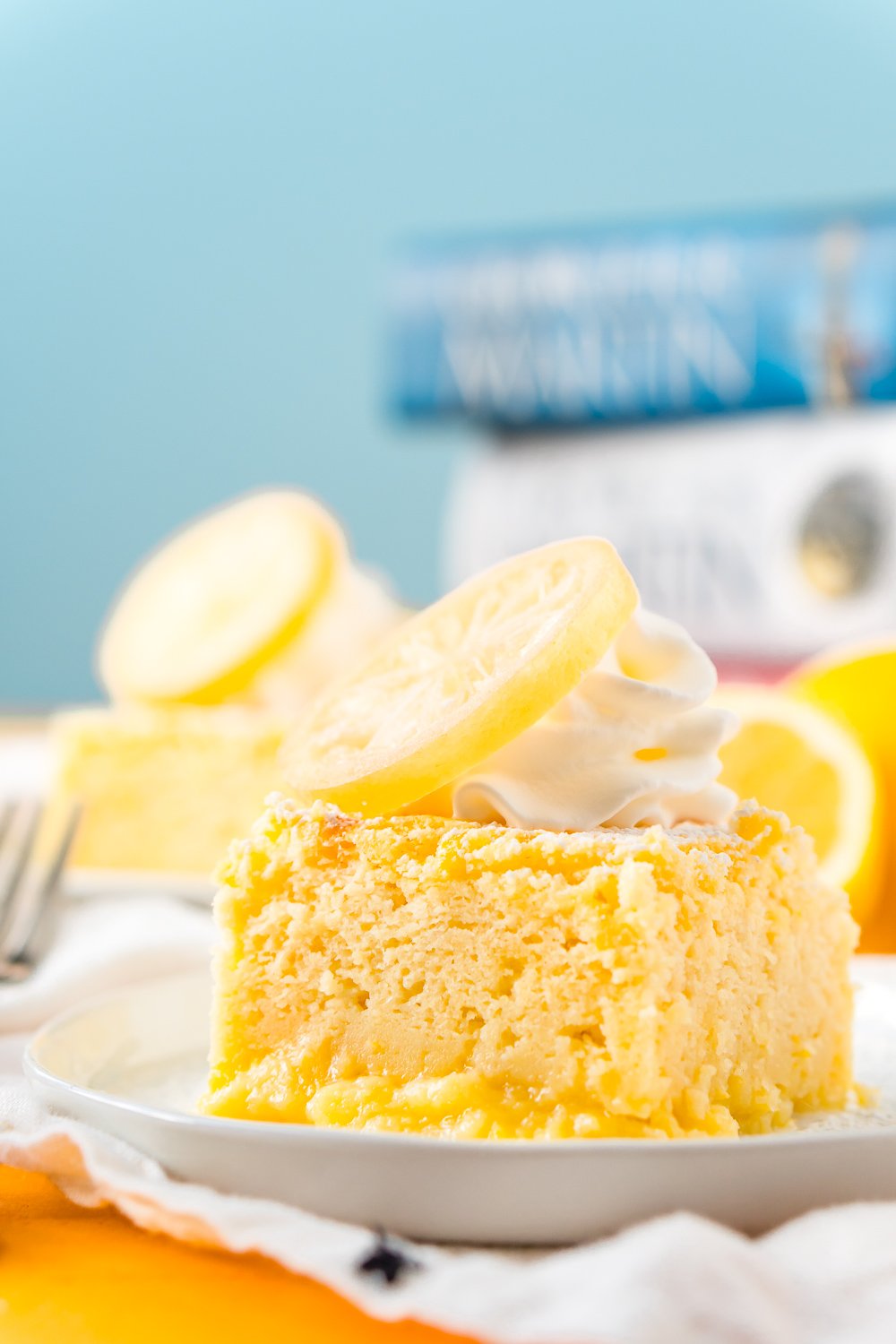 Piece of Lemon Magic Cake on a white plat with another slice of cake in the background and a fork next to it on the table.