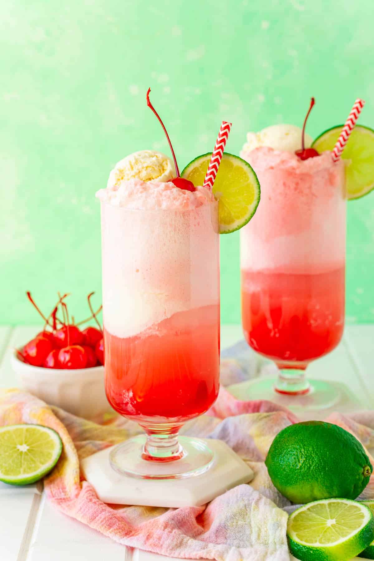 Two glasses of shirley temple floats on white coasters with limes and maraschino cherries around them.