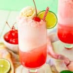 Shirley temple float on a white coaster with limes and maraschino cherries around it.