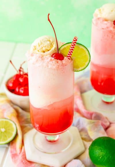 Shirley temple float on a white coaster with limes and maraschino cherries around it.