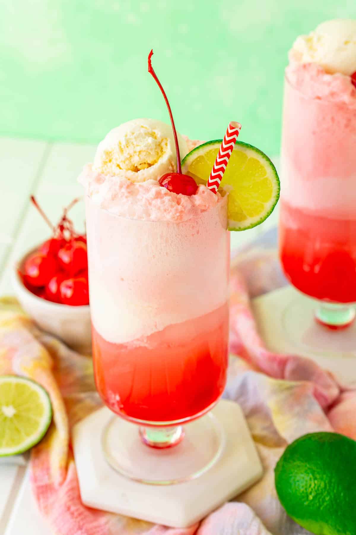 Shirley temple float on a white coaster with limes and maraschino cherries around it.