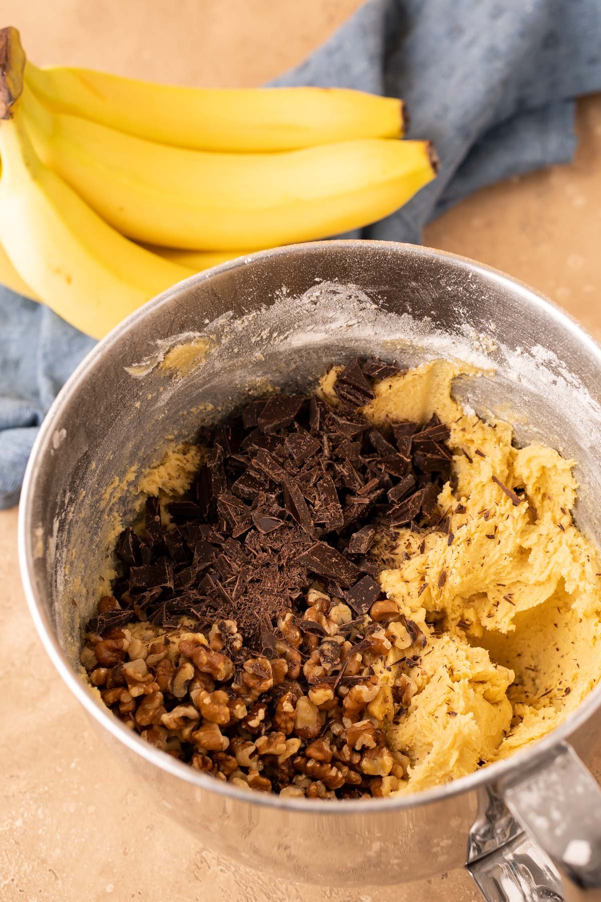 Walnut and chopped chocolate being added to a stand mixer bowl filled with cookie dough.