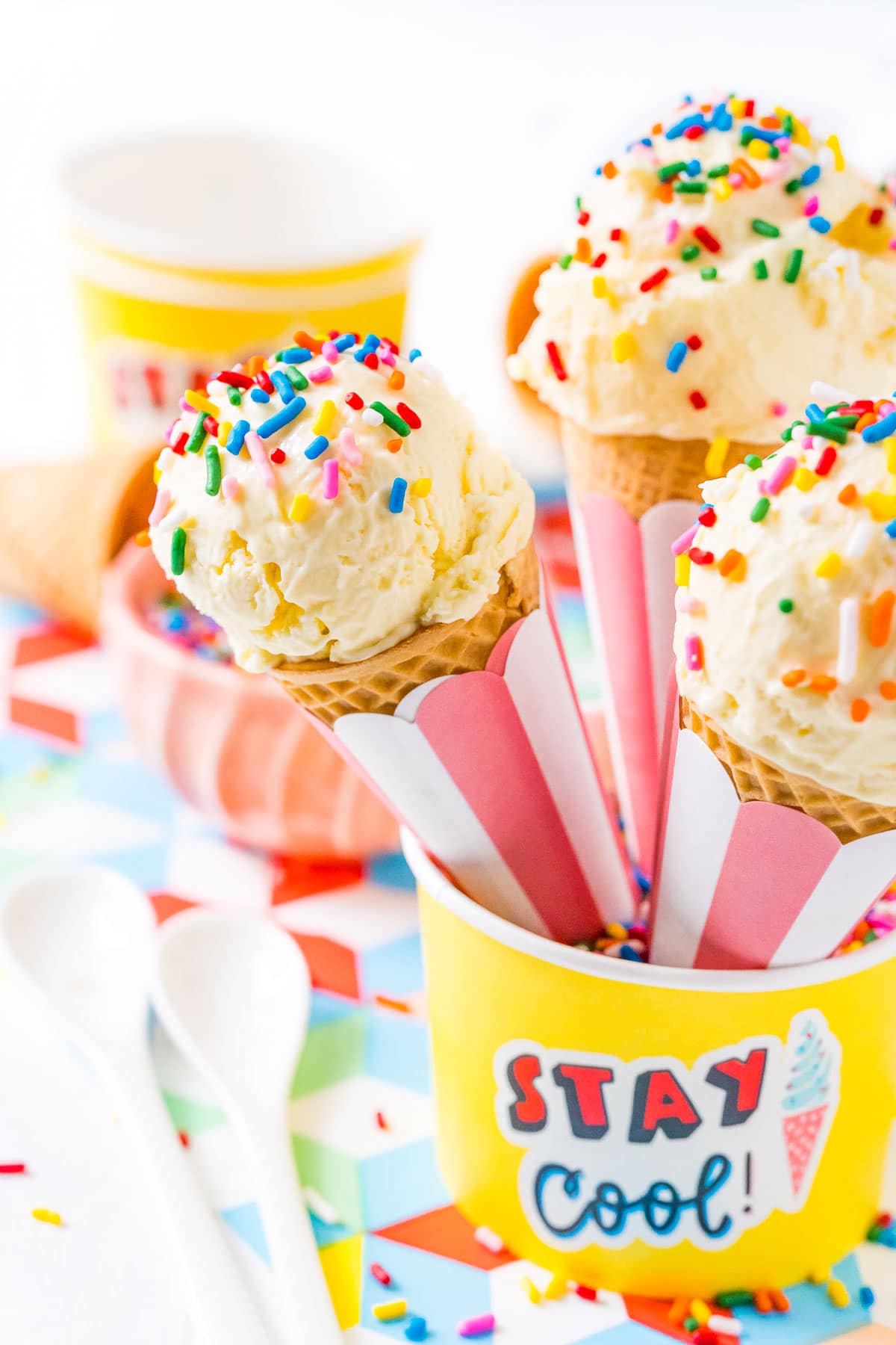 Three sugar cones with vanilla ice cream and sprinkles resting in a yellow bowl.