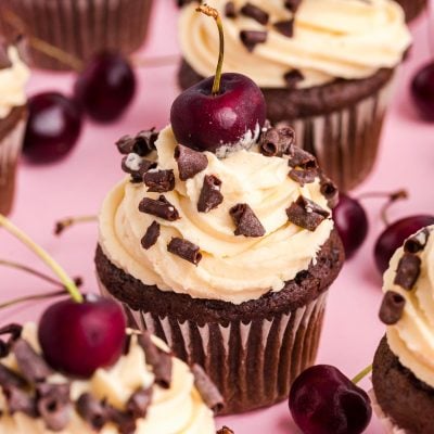 Black Forest Cupcakes on a pink surface surrounded by cherries.
