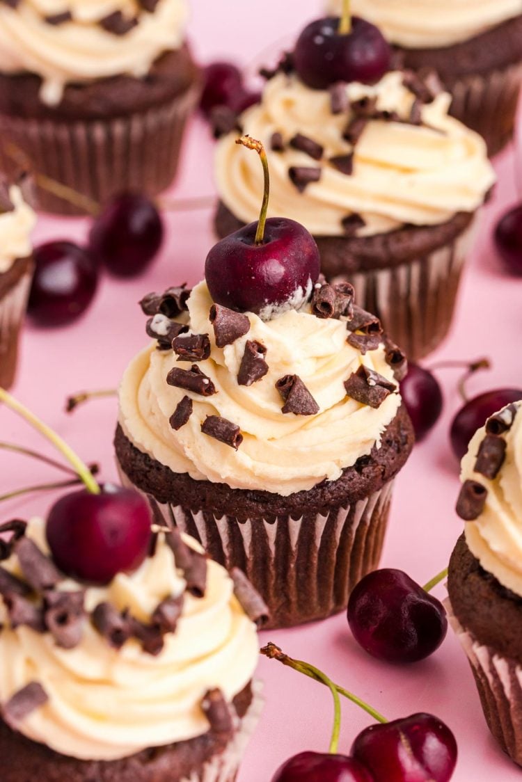 Black Forest Cupcakes on a pink surface surrounded by cherries.