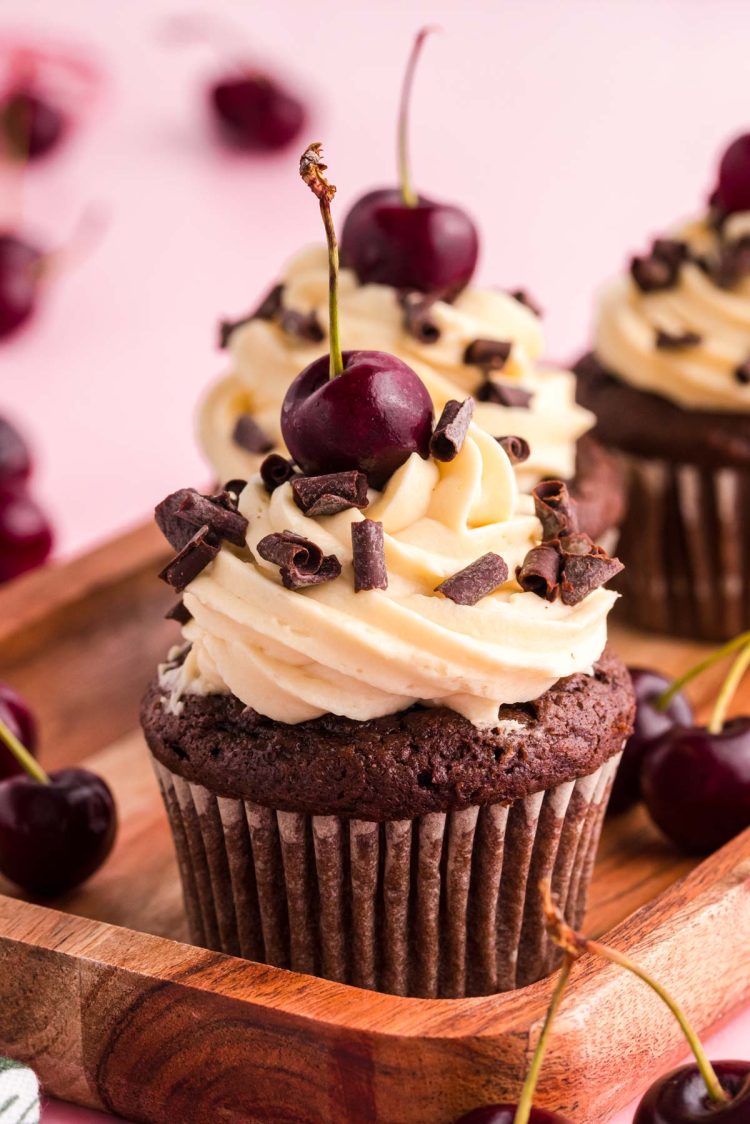 Close up photo of black forest cupcakes on a wooden tray on a pink surface.