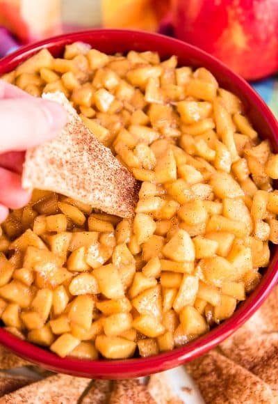 Woman's hand dipping a piece of pie crust into a red bowl filled with apple pie dip.