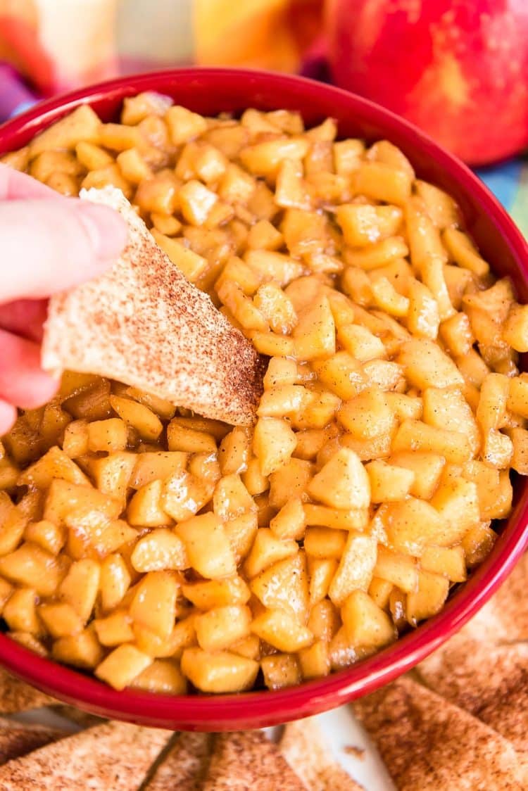 Woman's hand dipping a piece of pie crust into a red bowl filled with apple pie dip.