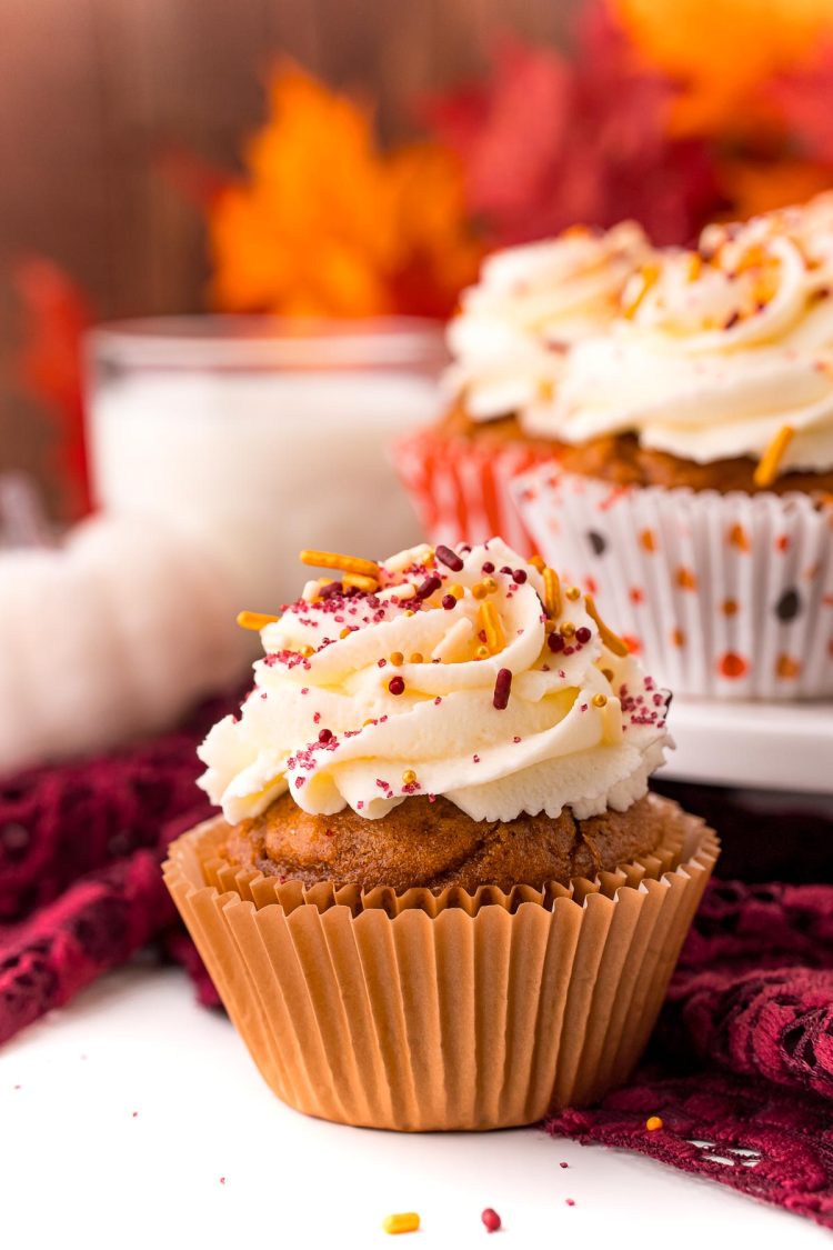 Close up photo of a pumpkin cupcake on a white table with a maroon napkin and more cupcakes and a glass of milk in the background.