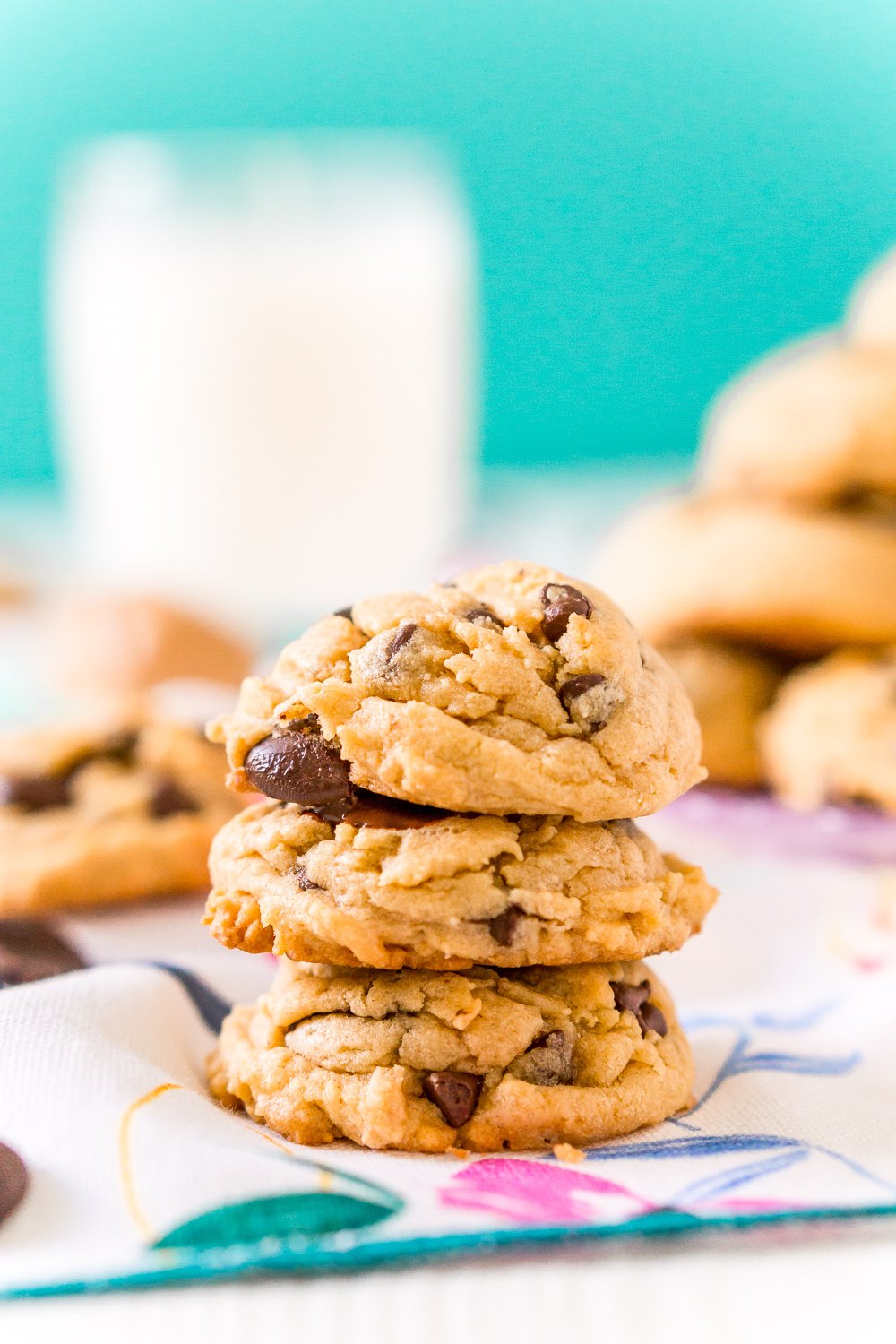 Three Peanut Butter Chocolate Chip Cookies stacked on top of each other with milk and more cookies in the background.