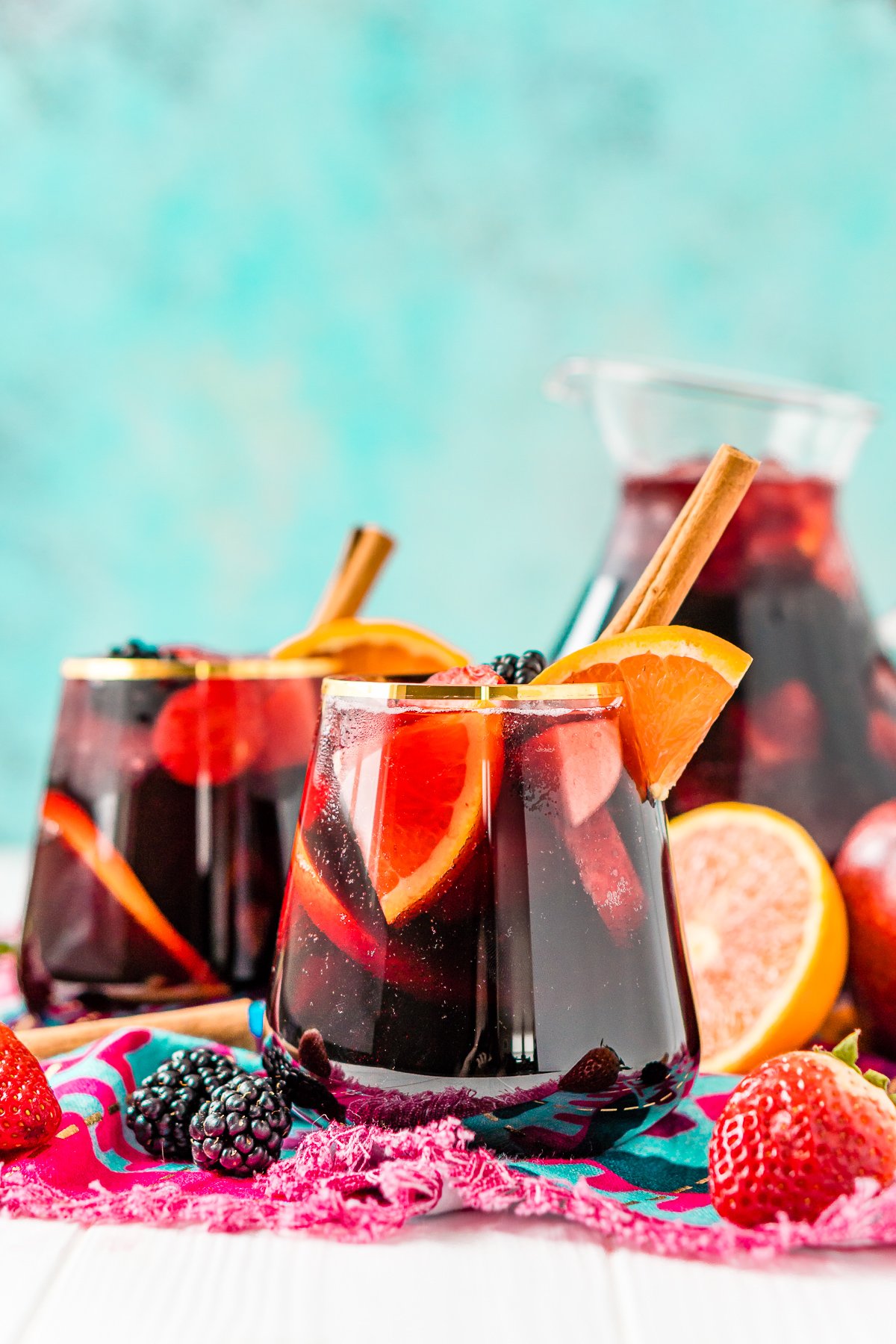 Glasses of red wine sangria in front of a full pitcher. Fresh fruit scattered around glasses. 