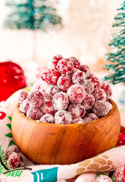 Close up photo of sugared cranberries in a wooden bowl surrounded by holiday decorations.