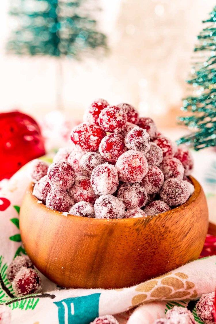 Close up photo of sugared cranberries in a wooden bowl surrounded by holiday decorations.