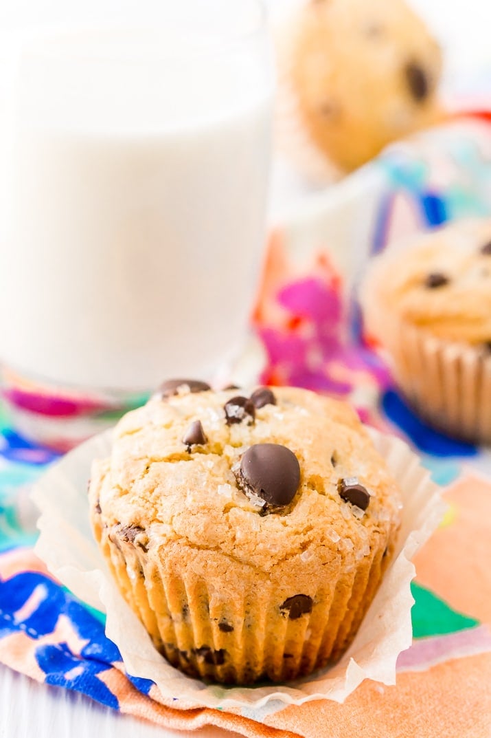 Close up photo of a chocolate chip muffins with a glass of milk and more muffins in the background.