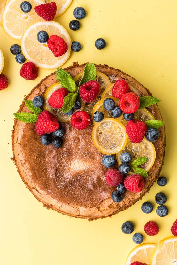 Overhead photo of lemon olive oil cake topped with berries and candied lemons on a yellow surface.