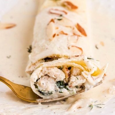 Close up photo of a spinach and chicken filled crepe with a gold fork taking a bite out of it from a white plate.