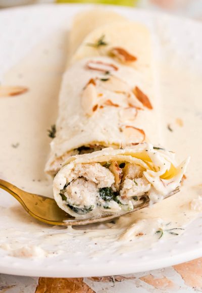 Close up photo of a spinach and chicken filled crepe with a gold fork taking a bite out of it from a white plate.